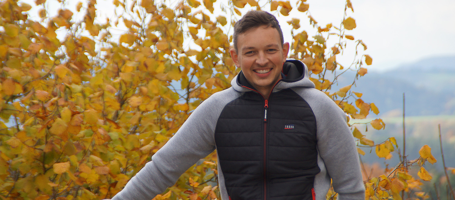 Maximilian Steindl steht vor einer Herbstlandschaft und lächelt in die Kamera.