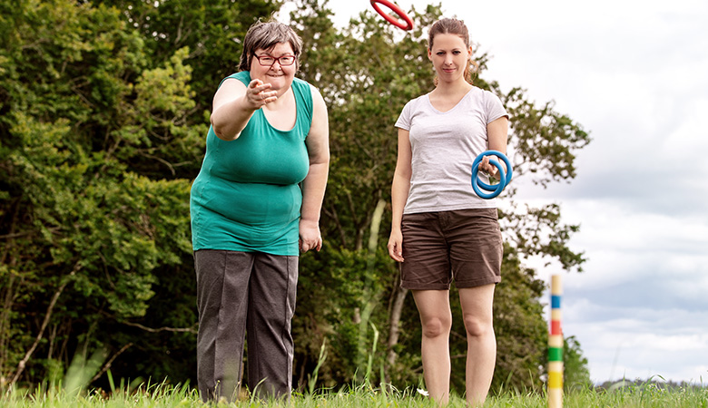  Eine Frau mit geistiger Behinderung spielt mit ihrer Betreuerin ein Ringwurfspiel auf einer Wiese.