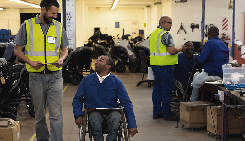 In einer Werkstatt geht ein Mann mit Warnweste neben einem Mann her, der im Rollstuhl sitzt und Arbeitskleidung trägt. Sie schauen sich lächelnd an. 