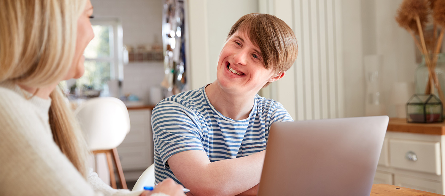 Eine Frau und ein junger Mann mit Down-Syndrom sitzen in einer Wohnung an einem Tisch. Sie lächeln sich an. Vor dem jungen Mann steht ein geöffneter Laptop.
