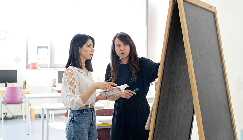 Yvonne Fritz steht mit einer Mitarbeiterin vor einem Whiteboard und zeigt darauf. Ihre Mitarbeiterin hält ein Buch in der Hand, sie selbst einen Stift.