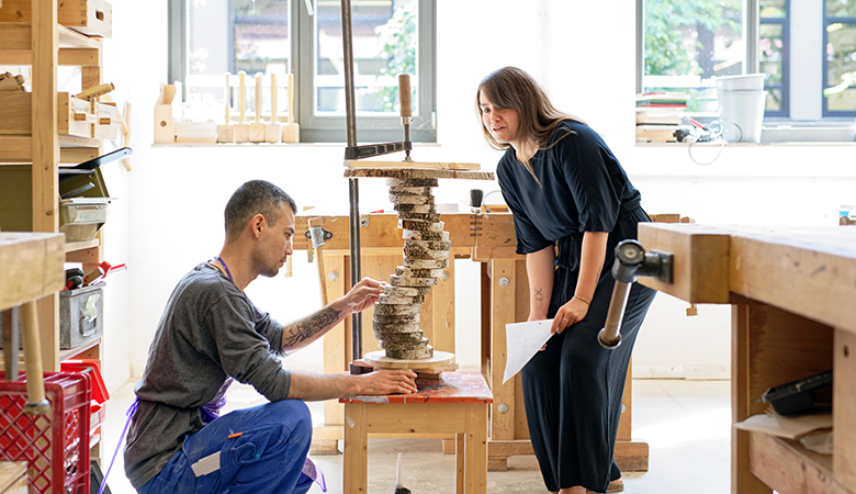 Yvonne Fitz steht mit einem jungen Mann in der Werkstatt. Er trägt Arbeitskleidung und kniet vor einer Holzkonstruktion. Yvonne Fritz begutachtet das Werk lächelnd und hält ein Blatt Papier in der Hand.