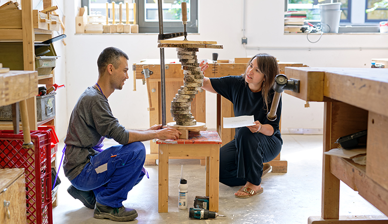 Yvonne Fitz kniet mit einem jungen Mann in der Werkstatt. Er trägt Arbeitskleidung und kniet vor einer Holzkonstruktion. Yvonne Fritz begutachtet das Werk lächelnd und hält ein Blatt Papier in der Hand. 