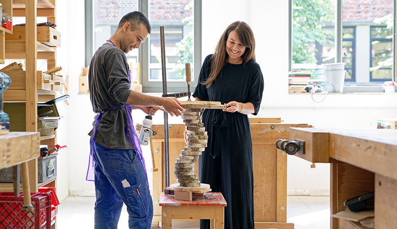 Yvonne Fitz steht mit einem jungen Mann in der Werkstatt. Er trägt Arbeitskleidung und arbeitet an einer Holzkonstruktion. Yvonne Fritz begutachtet das Werk lächelnd.
