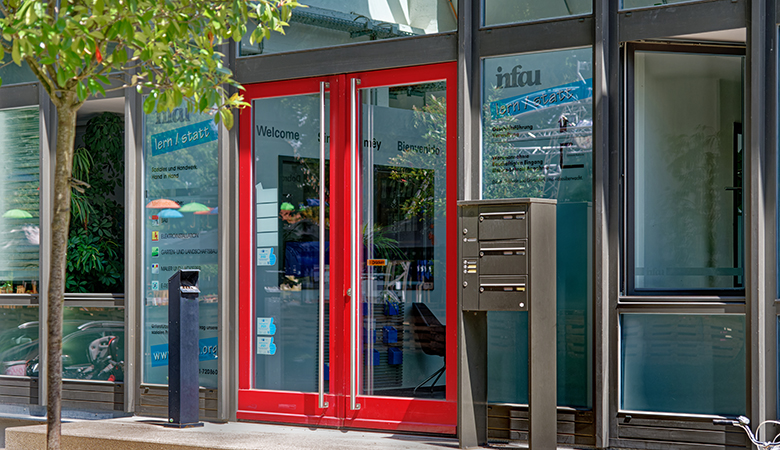 Das Eingang der infau-lern/statt GmbH mit einer roten Glastür. Davor steht ein Briefkasten. Die Scheiben neben der Tür sind mit Informationen beklebt.