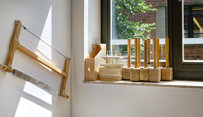Auf einem Fensterbrett stehen hölzerne Hammer und andere Bauelemente aus Holz. An der Wand daneben hängt eine Säge.