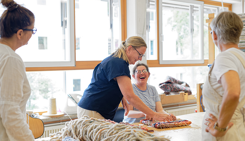 Vier Frauen sind in der Weberei, unter ihnen Kunigunde Frey. Auf dem Tisch liegt ein Teppich und Garn. Eine Frau sitzt am Tisch und arbeitet am Teppich, während Frau Frey ihr hilft. Die zwei weiteren Frauen stehen daneben. Sie lachen.