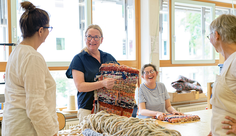 Vier Frauen sind in der Weberei. Eine von ihnen ist Kunigunde Frey. Sie hält einen Teppich in den Händen und zeigt ihn den anderen. Eine andere sitzt am Tisch mit einem weiteren Teppich. Die zwei weiteren Frauen stehen daneben. Alle lachen.