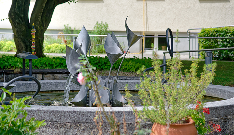 Ein Steinbrunnen im Außenbereich. Das Wasser fließt aus einer Metallkonstruktion.