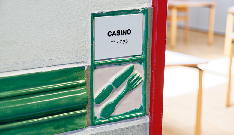 Zwei Fliesen sind an der Wand angebracht. Auf der oberen steht "Casino" in Buchstaben und Blindenschrift. Die Fliese darunter zeigt ein Messer und eine Gabel.