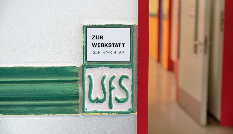Zwei Kacheln sind an einer Wand angebracht. Auf der oberen steht in Buchstaben und Blindenschrift "Zur Werkstatt". Auf der unteren Steht "WfS".