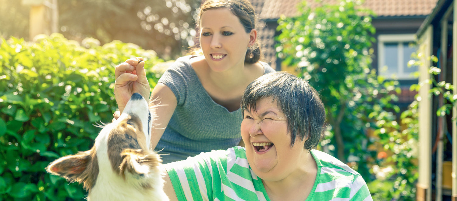 Zwei Frauen spielen mit einem Hund; eine der beiden hat eine geistige Behinderung.