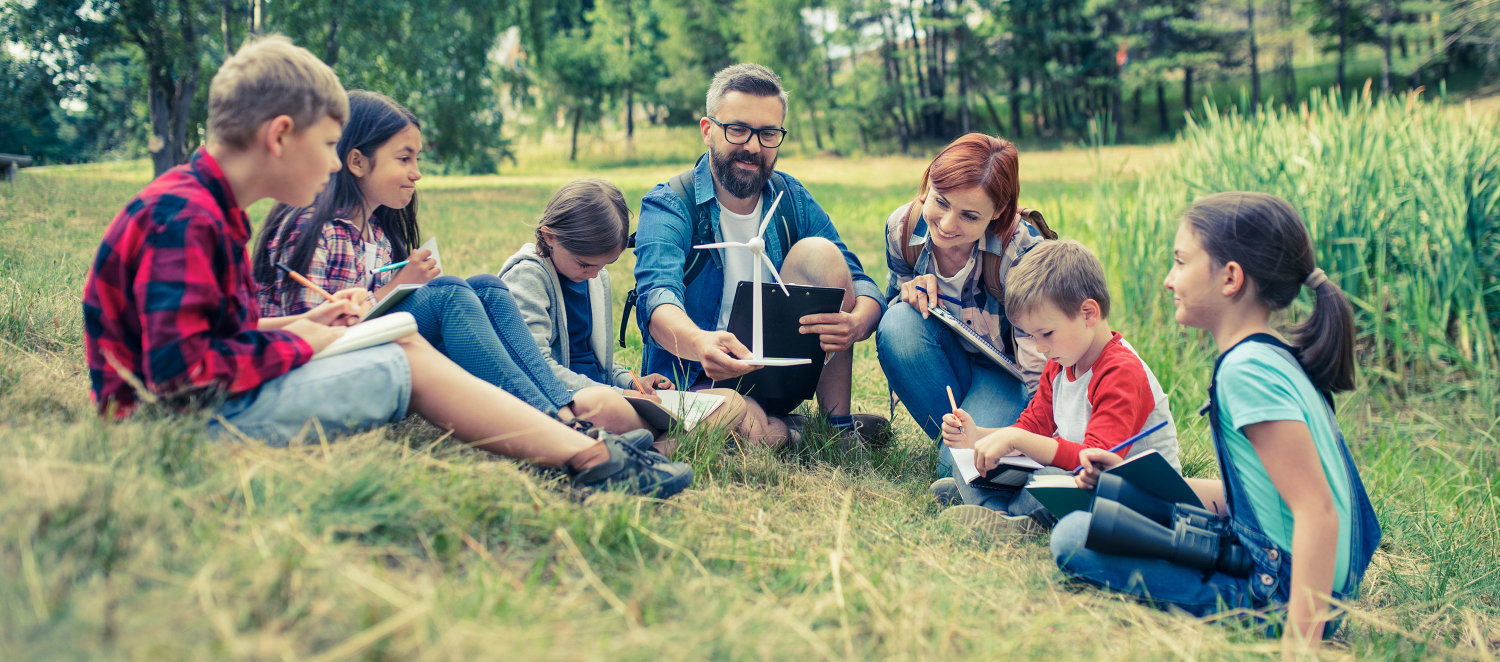 Erzieher und Erzieherin sitzen mit mehreren Kindern im Gras und erklären ihnen etwas.