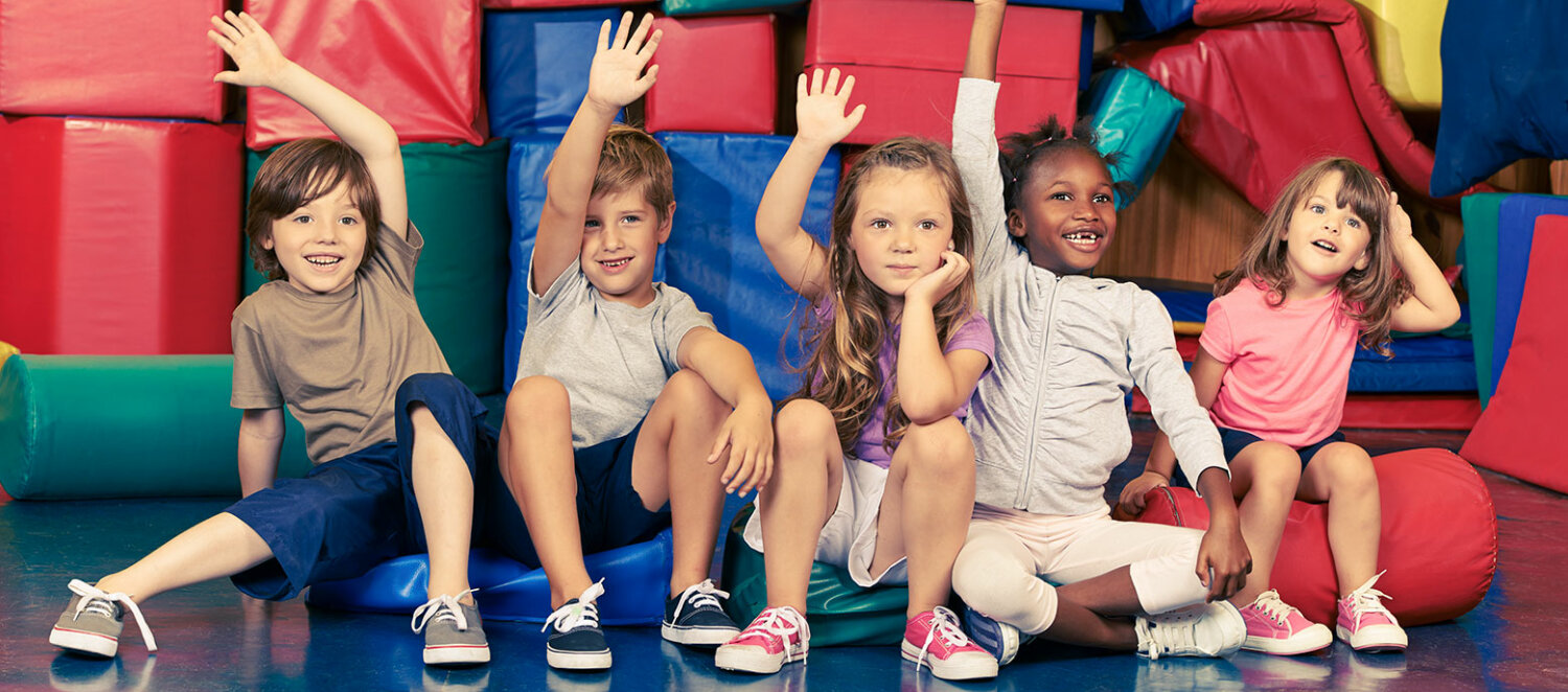 Szene im Kindergarten: Kinder sitzen vor Turnmatten auf dem Boden und halten die Hände hoch.