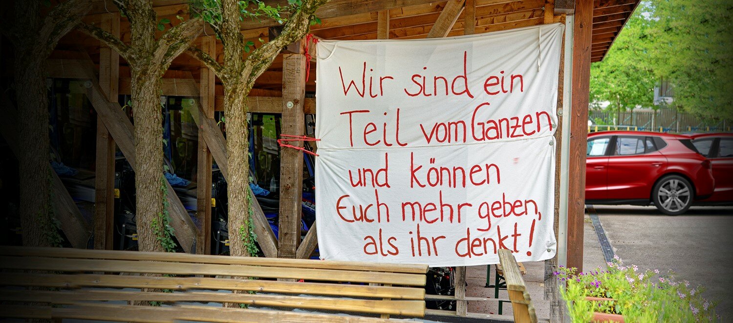 Ein Transparent mit der Aufschrift "Wir sind ein Teil vom Ganzen und können Euch mehr geben, als ihr denkt!" hängt an einem Zaun.