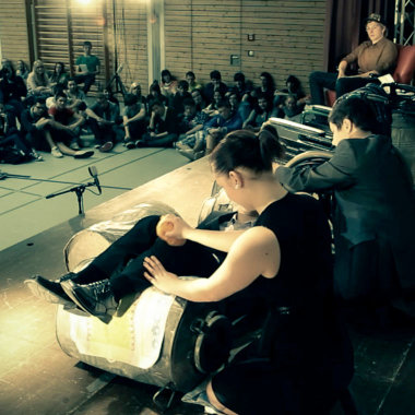 Szene in einer Schulturnhalle: Auf der Bühne sitzende Jugendliche werden von einem auf dem Boden sitzenden Publikum angesehen.
