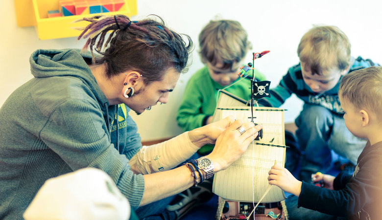 Szene im Kindergarten: Ein Erzieher und drei kleine Jungen spielen konzentriert mit einem Spielzeug-Piratenschiff.