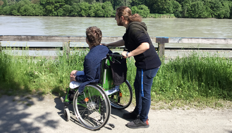 Zwei Männer blicken auf eine Flusslandschaft, einer von ihnen sitzt im Rollstuhl. 