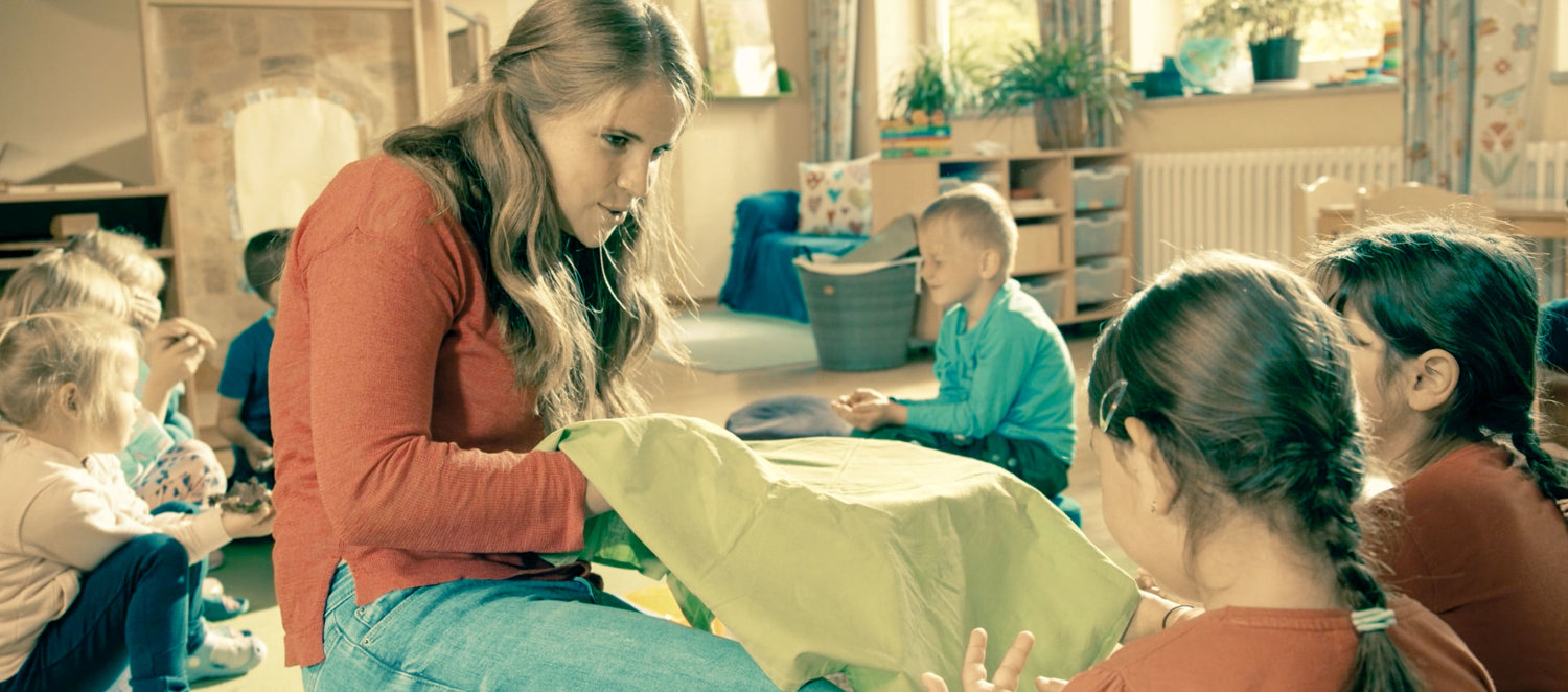 Szene im Kindergarten: Erzieherin versteckt etwas unter einem Tuch. Kinder raten, was es ist.