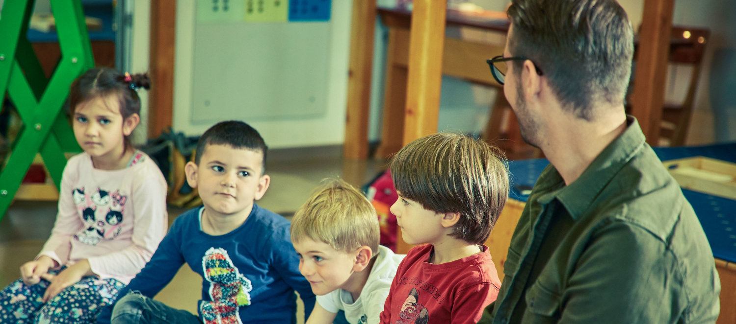 Szene im Kindergarten: Kinder und Erzieher sitzend auf dem Boden beim Morgenkreis.