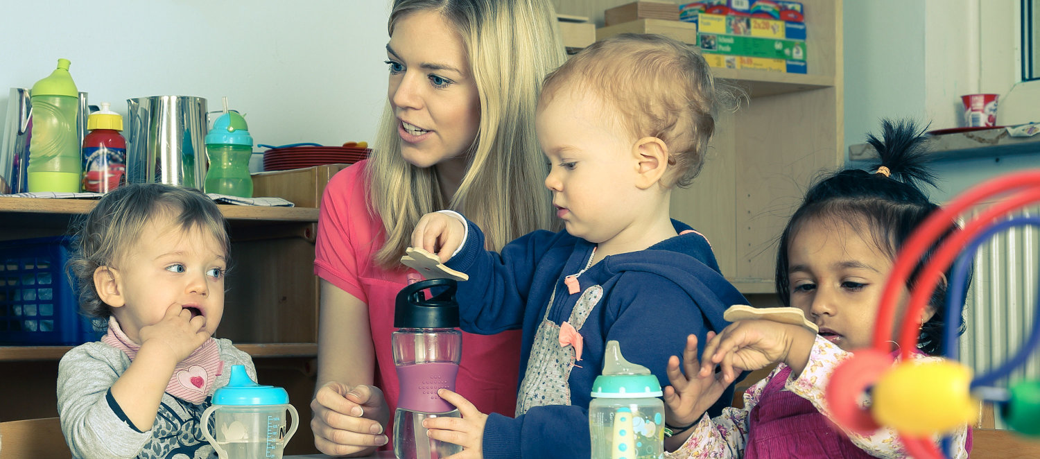 Szene im Kindergarten: Erzieherin mit Kleinkindern am Tisch beim Essen und Trinken.