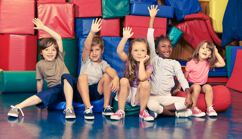 Szene im Kindergarten: Kinder sitzen vor Turnmatten auf dem Boden und halten die Hände hoch.