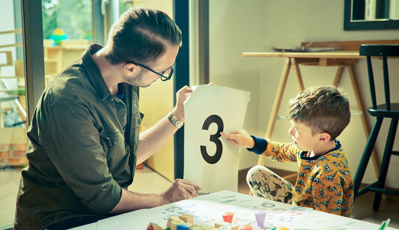 Szene im Kindergarten: Erzieher und Junge betrachten gemeinsam ein Blatt mit der Zahl Drei.