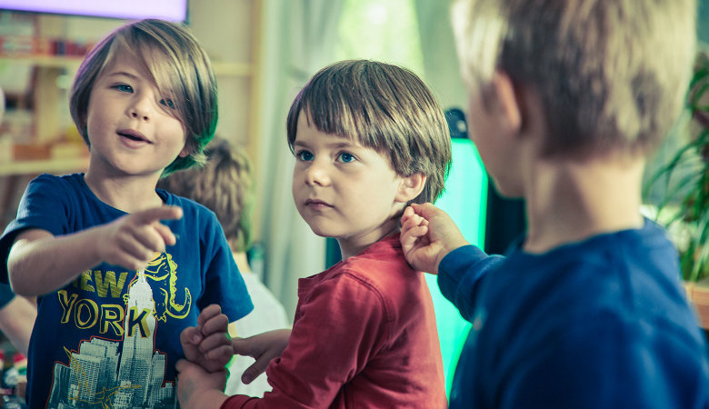 Szene im Kindergarten: Drei Kinder unterhalten sich. Einer zeigt mit dem Finger auf einen anderen.