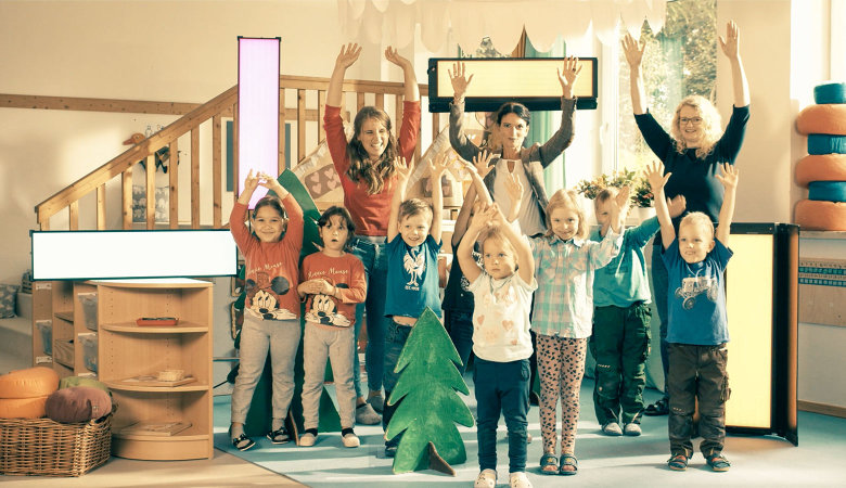 Szene im Kindergarten: Drei Erzieherinnen und sieben Kinder stehen lachend nebeneinander und strecken die Hände hoch.