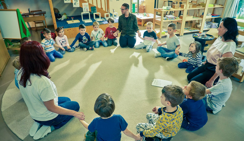 Szene im Kindergarten: Kinder und Erzieher knien im Kreis auf dem Boden und halten die Hände.