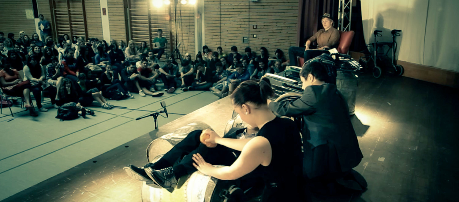 Jugendliche sitzen auf der Bühne einer Turnhalle, andere Jugendliche sitzen auf dem Boden und sehen zu.